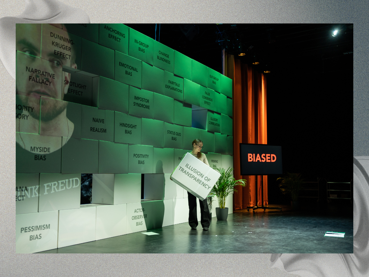 A person stands on a stage in front of a large wall made of stacked blocks, each labeled with different cognitive biases such as "Dunning-Kruger Effect," "Impostor Syndrome," and "Hindsight Bias." The person is holding a block labeled "Illusion of Transparency." A screen to the right displays the word "BIASED" in bold red letters. The scene is lit with green and orange lights, creating a dramatic atmosphere, with plants and stage curtains visible in the background.