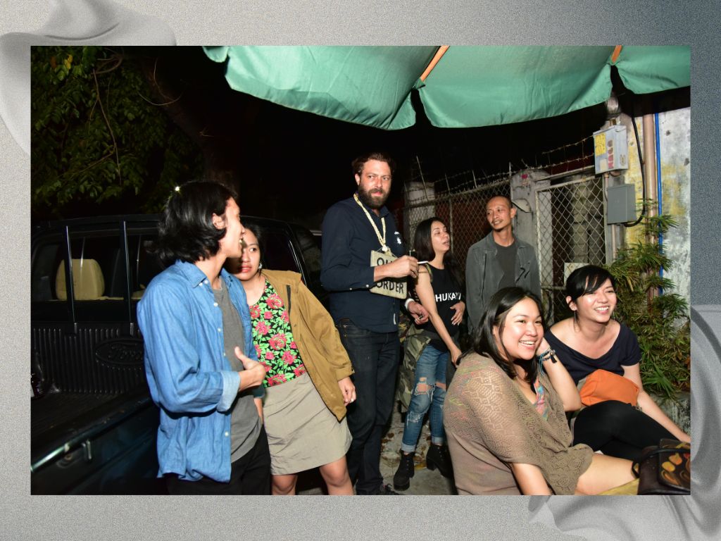 A casual outdoor gathering at night, featuring a group of seven people chatting and laughing under a green canopy. They are dressed in relaxed attire, with some seated and others standing near a parked vehicle. A fence and some greenery are visible in the background, creating an informal, friendly atmosphere.